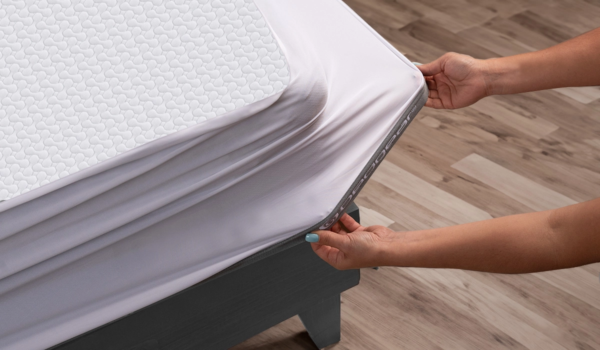 Person adjusting fitted sheet on textured mattress in minimalist, serene bedroom with wooden flooring.