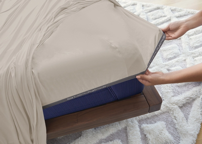Hands adjusting beige fitted sheet on blue mattress in a calm, modern bedroom.