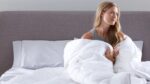 Young woman in serene bedroom, wrapped in cozy comforter, with calm, introspective expression.