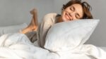 Young woman smiling in bed, embracing tranquility in a softly lit, cozy setting.