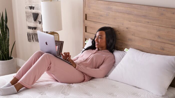 Woman working on a laptop in a cozy, stylish bedroom with wooden headboard.