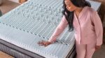 Woman testing light blue foam mattress for comfort in modern bedroom.