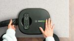 GhostBed massage device on rug, hands using controls, with remote for personalized comfort.