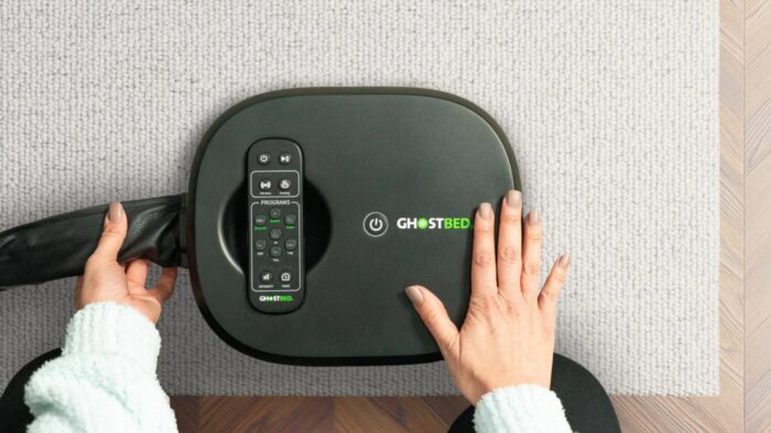 GhostBed massage device on rug, hands using controls, with remote for personalized comfort.