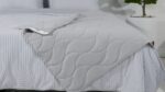Minimalist bed with gray quilt, striped sheets, pillows, and greenery for a tranquil atmosphere.