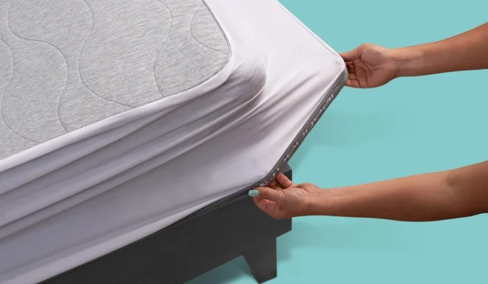 Hands fitting white mattress protector on gray mattress with teal background for hygiene and comfort.